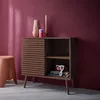 A wooden cabinet with a ribbed sliding door is placed on a grey floor against a dark red wall. Decorative items such as vases and candles are on top of the cabinet. A pink stool is next to the cabinet. Anteprima dell’immagine prodotto
