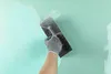 A person's hand wearing a grey glove is applying a light blue substance to a green wall with a trowel. Produktbild-Vorschau