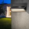 A black outdoor wall light is mounted on a textured gray wall, illuminating a garden and house entrance at dusk. Produktbild-Vorschau