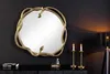 A decorative mirror with a snake-shaped frame is displayed on a dark wall above a black dresser. Two brass candlesticks with lit candles and a small perfume bottle are placed on the dresser. Produktbild-Vorschau