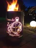 A fire pit with a clock design is lit at night, with a glowing orb light in the background. Podgląd zdjęcia produktu