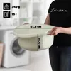 A person is holding a cream-colored plastic basin in front of a blurred background of a laundry room with a washing machine. Produktbild-Vorschau