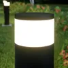 A close-up of a black bollard light with a white illuminated panel, set in a garden with green grass and blurred red flowers in the background. Produktbild-Vorschau