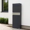 A modern anthracite mailbox with a silver accent strip is mounted on a white exterior wall next to a window. The mailbox is positioned on a paved walkway with green bushes in the background. Voir les images du produit