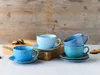 A collection of blue and teal ceramic cups and saucers are arranged on a light-colored surface, with a wooden background and some cookies on a wooden board. Náhľad obrázku produktu