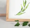A close-up of a wooden picture frame with a watercolor painting of green leaves inside, and a real branch with leaves in the foreground. Produktbild-Vorschau