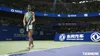A female tennis player stands on a tennis court in a stadium, holding a racket. The background shows spectators and advertising banners. Produktbild-Vorschau
