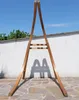 A wooden hammock stand is set up outdoors on a stone patio, with a white wall and green trees in the background under a clear blue sky. Produktbild-Vorschau