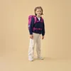 A young girl wearing a backpack stands against a plain background. Produktbild-Vorschau
