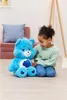 A young girl is sitting on the floor next to a large blue plush Care Bear. Produktbild-Vorschau