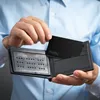 A person is holding a black wallet open and placing a black card into it. Produktbild-Vorschau
