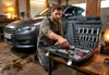 A man in work clothes is kneeling next to a car, opening a large toolbox filled with tools. Produktbild-Vorschau