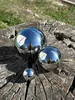 Three polished stainless steel garden spheres of different sizes are placed on a weathered wooden stump, with a person's reflection visible in the spheres. Náhled obrázku produktu