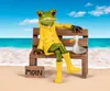 A frog figurine wearing a yellow raincoat and boots sits on a wooden bench on a beach, with a seagull figurine beside it. Produktbild-Vorschau