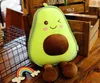 A plush avocado toy sitting on a wooden surface with wooden crates in the background. Produktbild-Vorschau