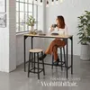 A woman sits at a high table with two bar stools, holding a mug. Náhled obrázku produktu