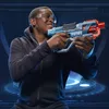A young person wearing safety glasses and a dark hoodie holds a blue and red toy gun. The background is a dark blue grid with glowing blue circular graphics. Anteprima dell’immagine prodotto