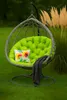 A hanging egg chair with a green cushion, a floral pillow, and a dark gray blanket, set on a green lawn with tall ornamental grasses in the background. Voir les images du produit