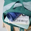 A close-up view of a teal tote bag with its zipper partially open, revealing a blue cup, a book, and a light blue laptop inside. The bag is placed on a wooden stool. Voir les images du produit