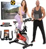 A man and a woman are posing with an indoor cycling bike, a large screen displaying a virtual cycling course, and accessories like a tablet and heart rate monitor. Produktbild-Vorschau