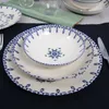 A set of white ceramic plates and bowls with blue decorative patterns, arranged on a dark blue placemat. Produktbild-Vorschau