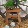 A wooden stool with a plant on it, placed on straw, with a large wagon wheel in the background. Náhled obrázku produktu