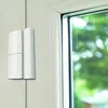 A white magnetic contact sensor attached to a door or window frame, next to a window. Produktbild-Vorschau