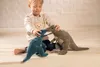 A young boy is playing with two stuffed dinosaur toys on a wooden floor in front of a brick wall. Produktbild-Vorschau