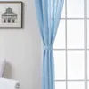 A light blue curtain is tied back with a fabric band, hanging in front of a window with white mullions. A framed picture of a building is visible on the left wall. Produktbild-Vorschau