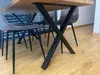 A close-up shot of black metal X-shaped table legs under a wooden tabletop, with gray chairs visible in the background. Anteprima dell’immagine prodotto