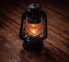 A black kerosene lantern with a lit wick sits on a wooden surface. Náhľad obrázku produktu