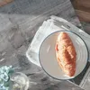 A croissant on a plate, placed on a newspaper, with a small vase of flowers in the foreground. Produktbild-Vorschau