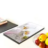 A glass cutting board with an orchid image is placed on a white surface next to a stack of books and a bowl of fruit. Voir les images du produit