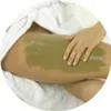 A person's hand is applying a green mud mask to a thigh, with white fabric visible. Voir les images du produit