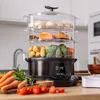 A multi-tiered food steamer filled with various vegetables and dumplings, placed on a kitchen counter with fresh produce in the foreground. Náhled obrázku produktu