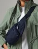 A person wearing a green jacket and a white t-shirt, with a navy blue fanny pack worn across their chest. Anteprima dell’immagine prodotto