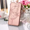 A rose gold flip phone case with a butterfly embossed design is displayed on a reflective surface, with decorative elements like beads, a teacup, and flowers in the background. Produktbild-Vorschau