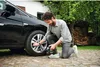 A man is kneeling next to a car, using a portable air compressor to inflate the tire. Produktbild-Vorschau