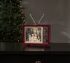 A decorative retro television with a Christmas scene inside, placed on a dark surface with a Christmas tree in the background. Podgląd zdjęcia produktu