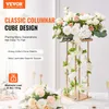 A gold columnar cube stand decorated with white flowers and green leaves, placed on a table with other floral arrangements. Voir les images du produit