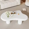 A white, irregularly shaped coffee table with a vase of flowers, books, and candles on it, placed in a living room setting with a white sofa. Produktbild-Vorschau