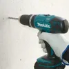 A hand wearing a white glove is holding a teal and black Makita drill, drilling a hole into a textured white wall. Produktbild-Vorschau