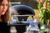 A woman is partially visible in the foreground, looking down at a black gas pizza oven on a table. The oven has wooden legs and a silver knob. In the background, there are pink flowers and a glass pitcher. Produktbild-Vorschau