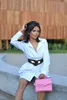 A woman is sitting on a concrete ledge outdoors, wearing a white shirt dress, a black belt, and holding a pink handbag. Produktbild-Vorschau