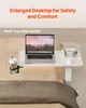 A white adjustable overbed table with a laptop, headphones, and a cup holder is shown on a bed. Produktbild-Vorschau