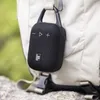 A black portable speaker with a carabiner clip is attached to a beige backpack, outdoors. Voir les images du produit