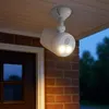A white battery-powered outdoor spotlight with motion sensor is mounted on a white ceiling under a porch, illuminating a brick wall and a dark wooden door. Produktbild-Vorschau