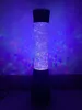 A tall, cylindrical glitter lamp with a black base and top, illuminated with blue and multicolor lights, casting colorful patterns on a purple background. Anteprima dell’immagine prodotto