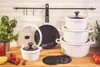 A collection of white cookware with black accents, including pots, pans, and lids, arranged on a wooden countertop with some fresh produce and greenery in the background. Anteprima dell’immagine prodotto