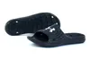 A pair of black Under Armour slides shown from a slightly elevated angle against a white background. One slide is shown upside down, revealing its sole, while the other is right-side up. Anteprima dell’immagine prodotto